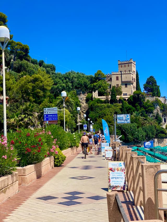 A sunny day on a picturesque Italian beach promenade, with vibrant blue skies, lush greenery, and historic architecture in the background