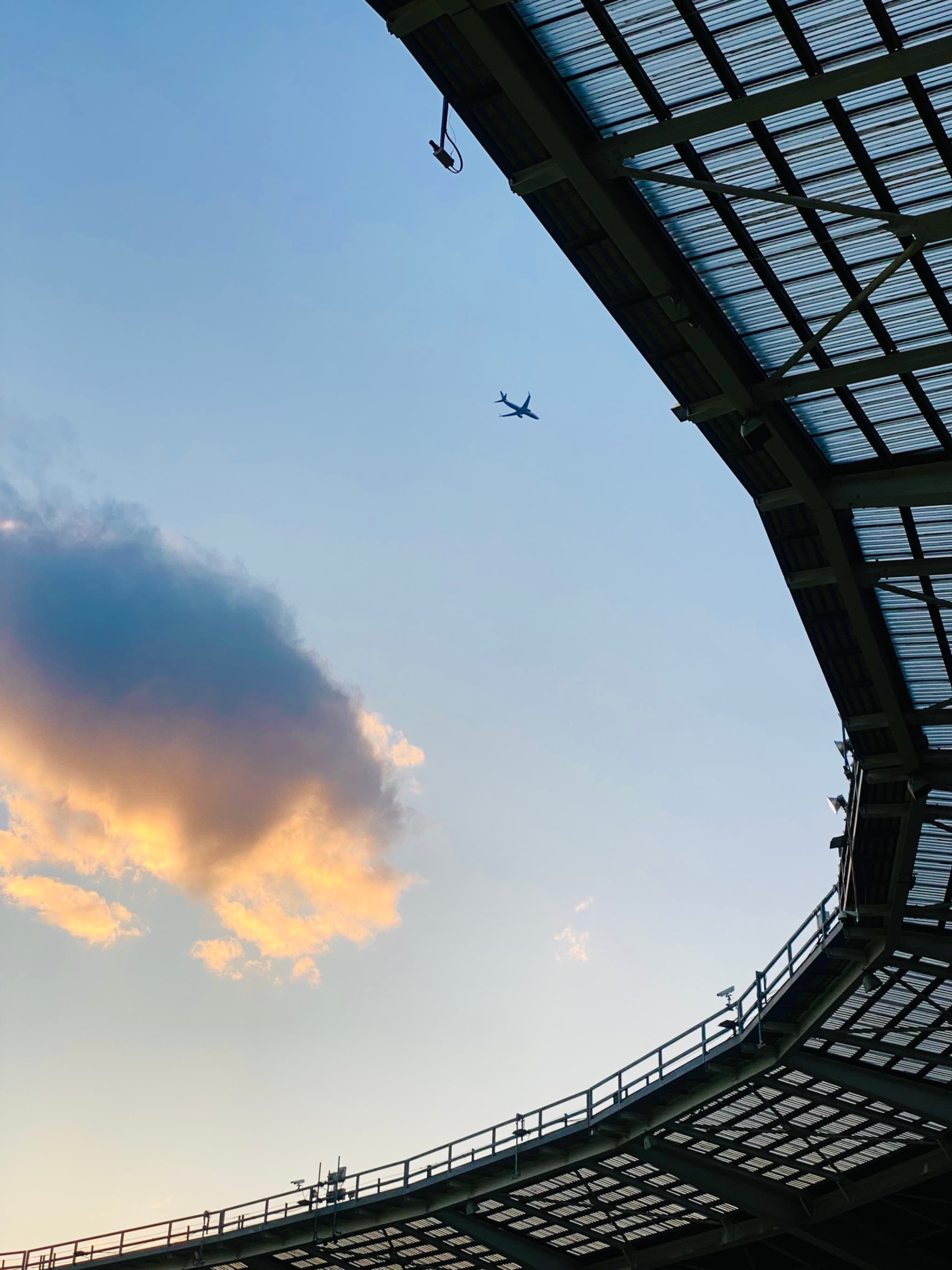 A serene sunset sky meets the underside of a grand metal structure, with a plane soaring overhead and a dramatic cloud adding depth