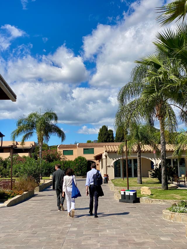 Three people stroll under a bright blue sky with fluffy clouds, surrounded by lush palm trees at a luxurious resort