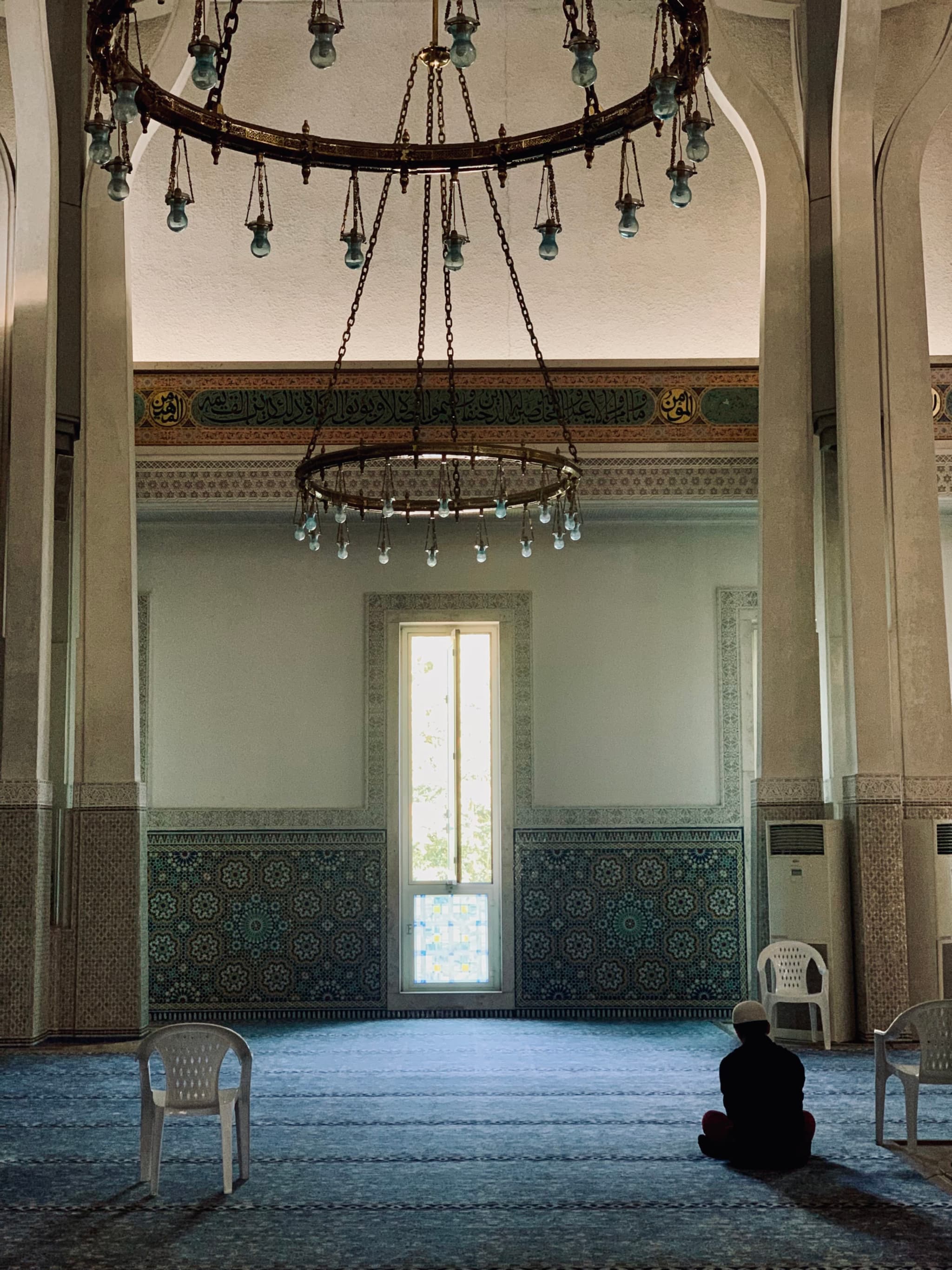 An Islamic prayer room with ornate tiles, grand chandelier, and pillars, lit by natural light filtering through a glass door, creating a serene, contemplative atmosphere