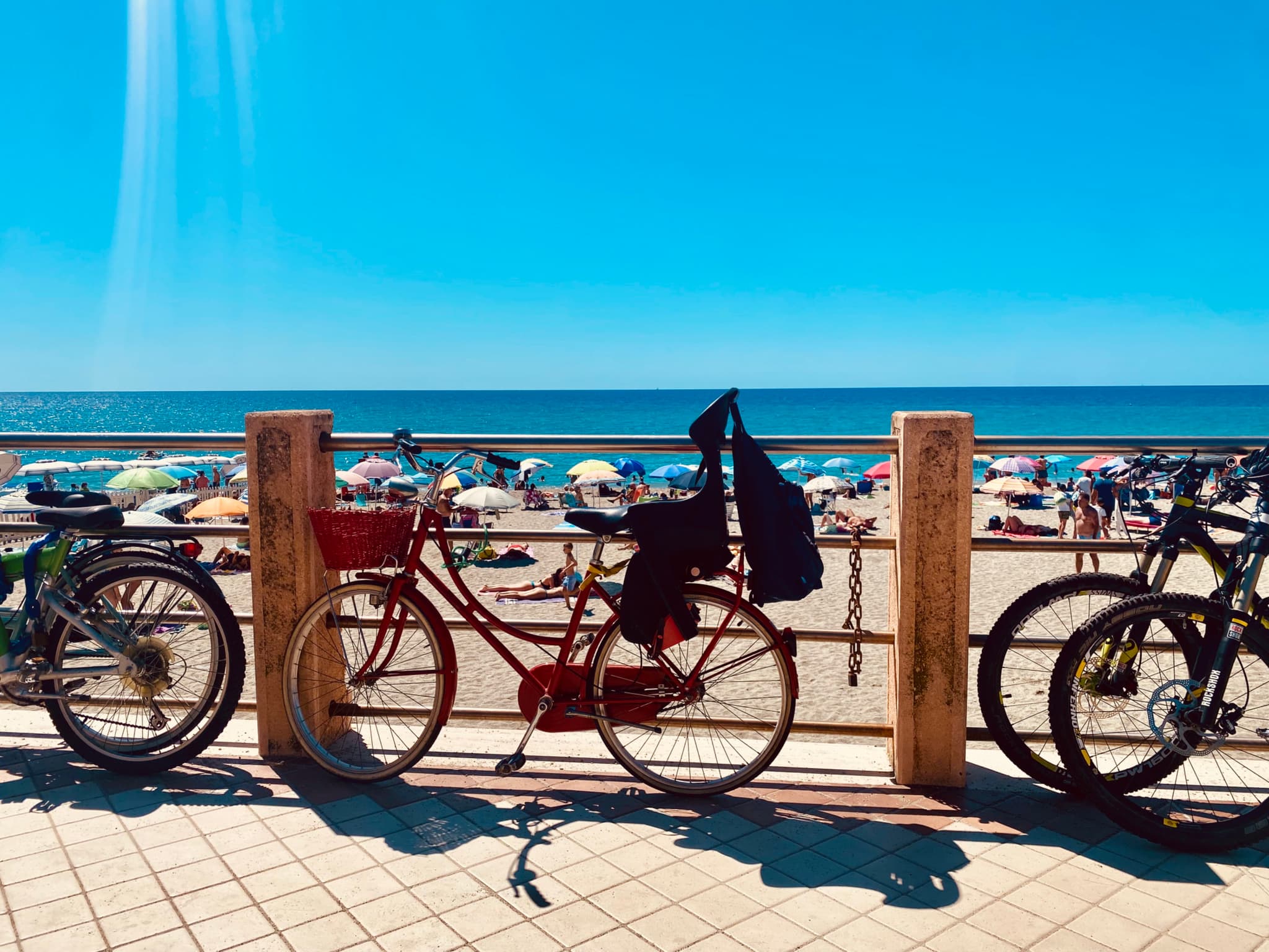 A vibrant beach scene with colorful bicycles under bright sunlight, figures lounging on the sand, and clear blue skies
