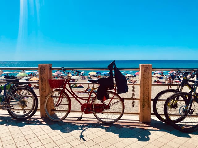 A vibrant beach scene with colorful bicycles under bright sunlight, figures lounging on the sand, and clear blue skies