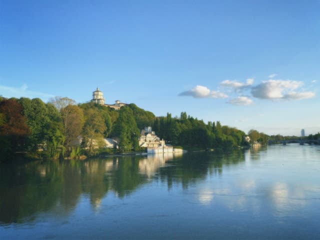 A serene lakeside scene with autumn foliage, a picturesque bridge, and a castle or tower in the distance, under a partly cloudy sky
