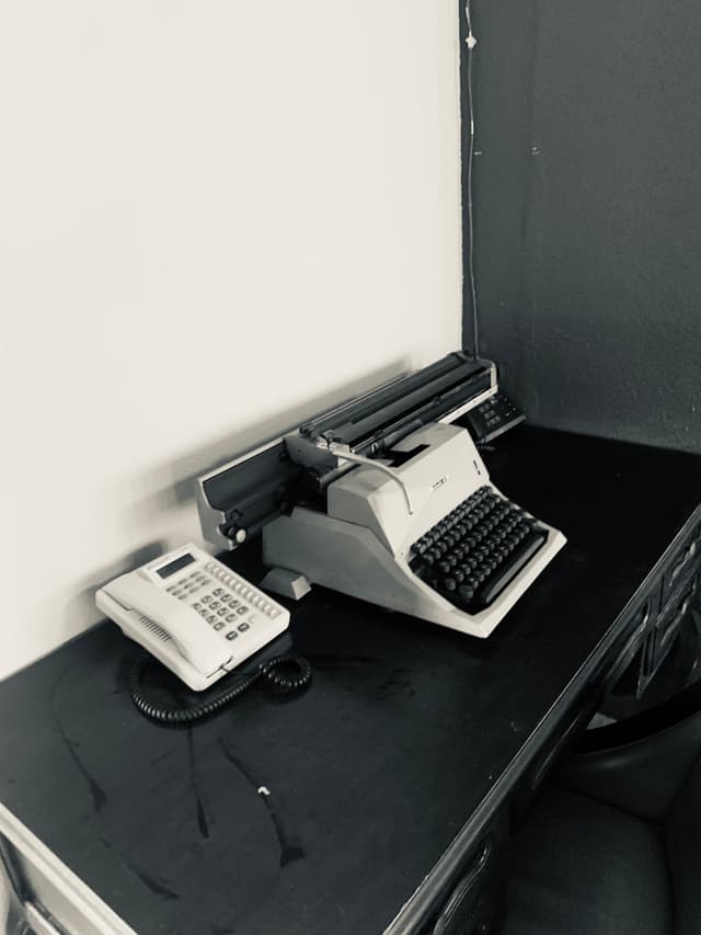 A vintage black-and-white typing scene: a typewriter on a black desk against a stark white wall, evoking a nostalgic office atmosphere