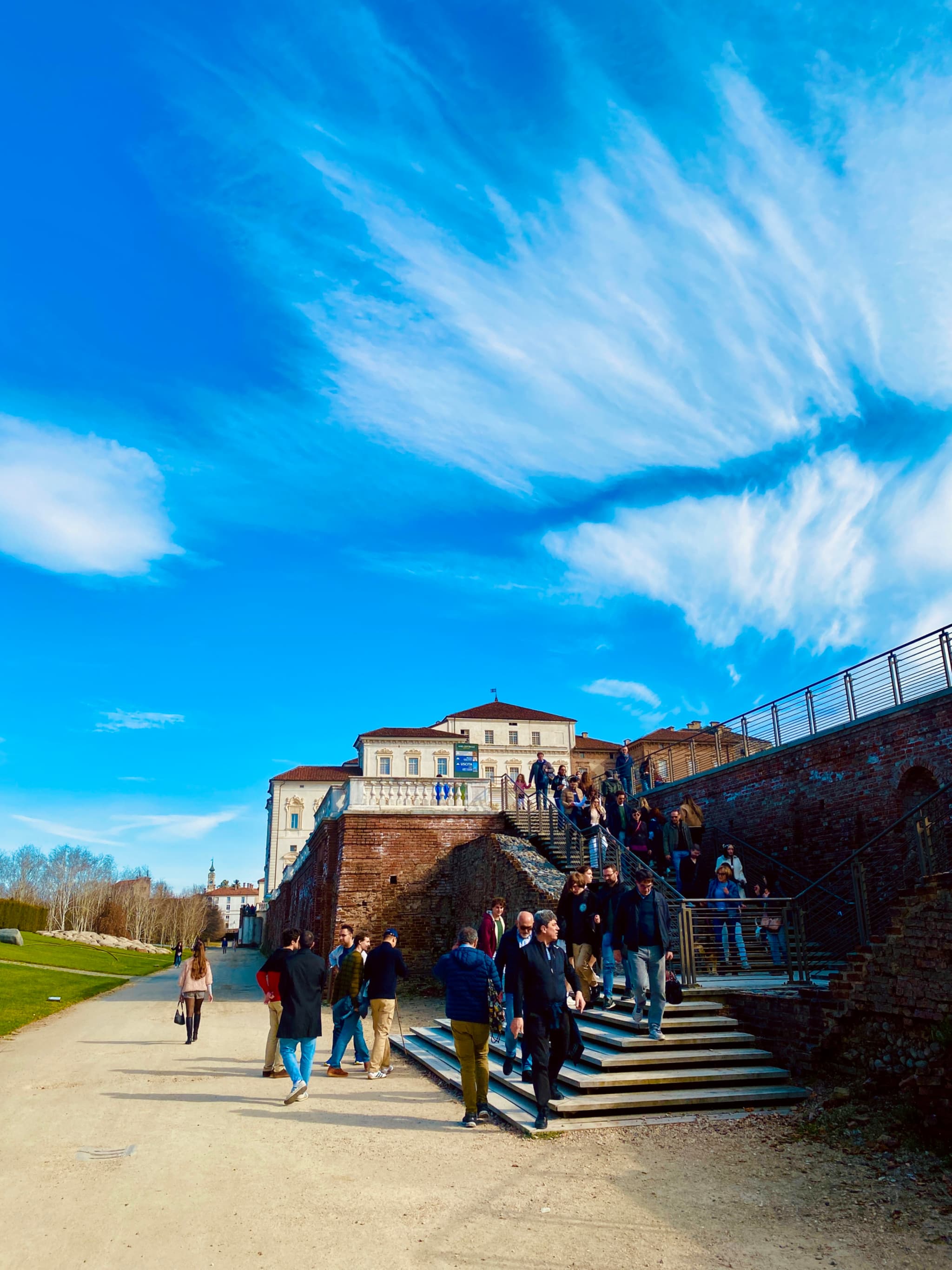A vibrant daytime scene at a grand, historic castle with people exploring the grand staircase, under a bright blue sky dotted with wispy clouds