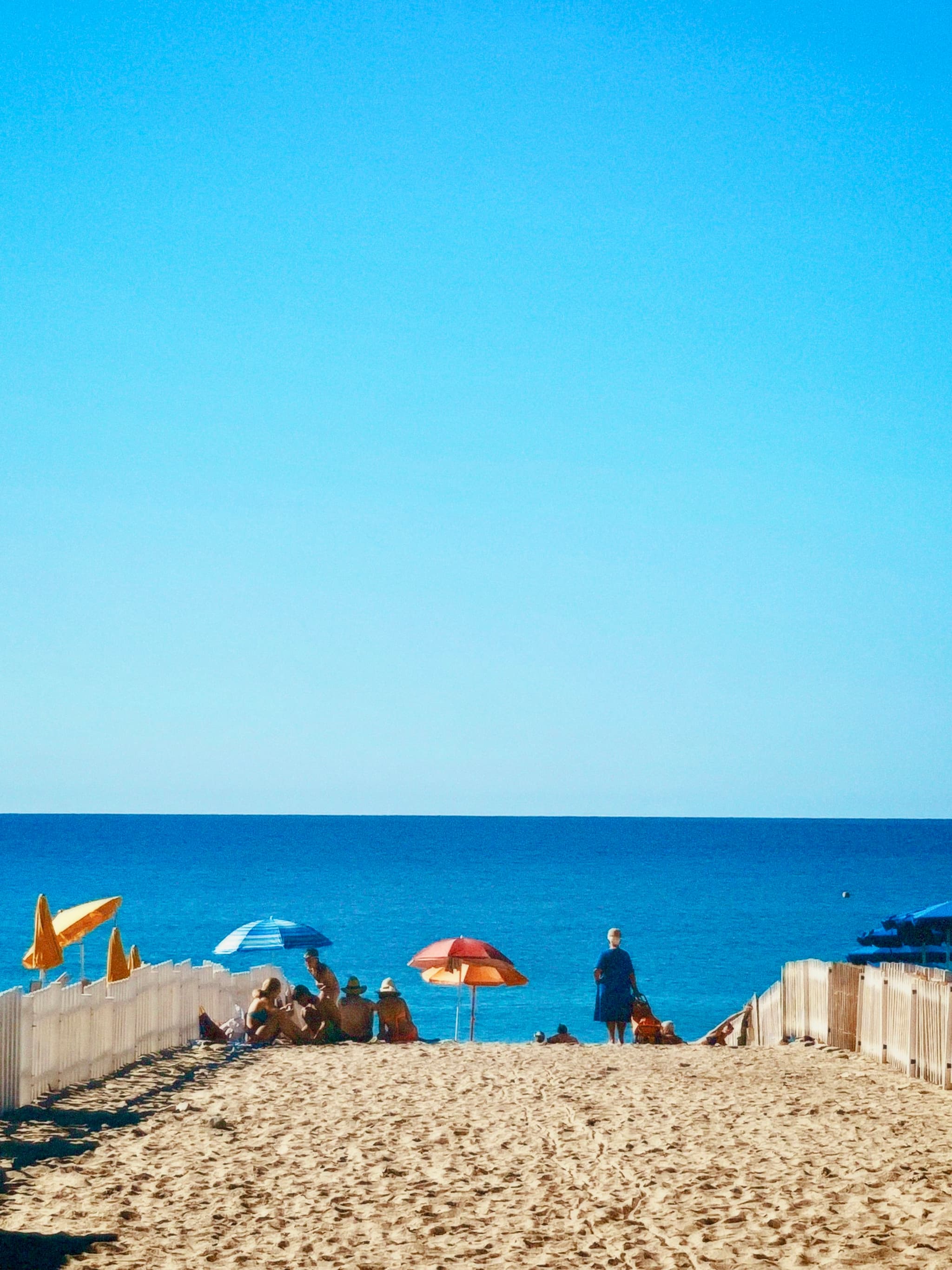 A sunny beach scene with colorful umbrellas, sandy paths, and a clear blue sky, evoking a relaxed, summery vibe