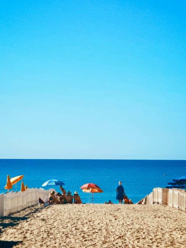 A sunny beach scene with colorful umbrellas, sandy paths, and a clear blue sky, evoking a relaxed, summery vibe