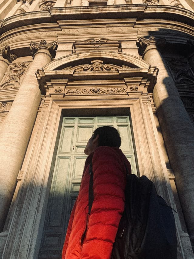 A woman in a red jacket stands before a grand, sunlit church entrance, casting long shadows and evoking a serene, contemplative mood
