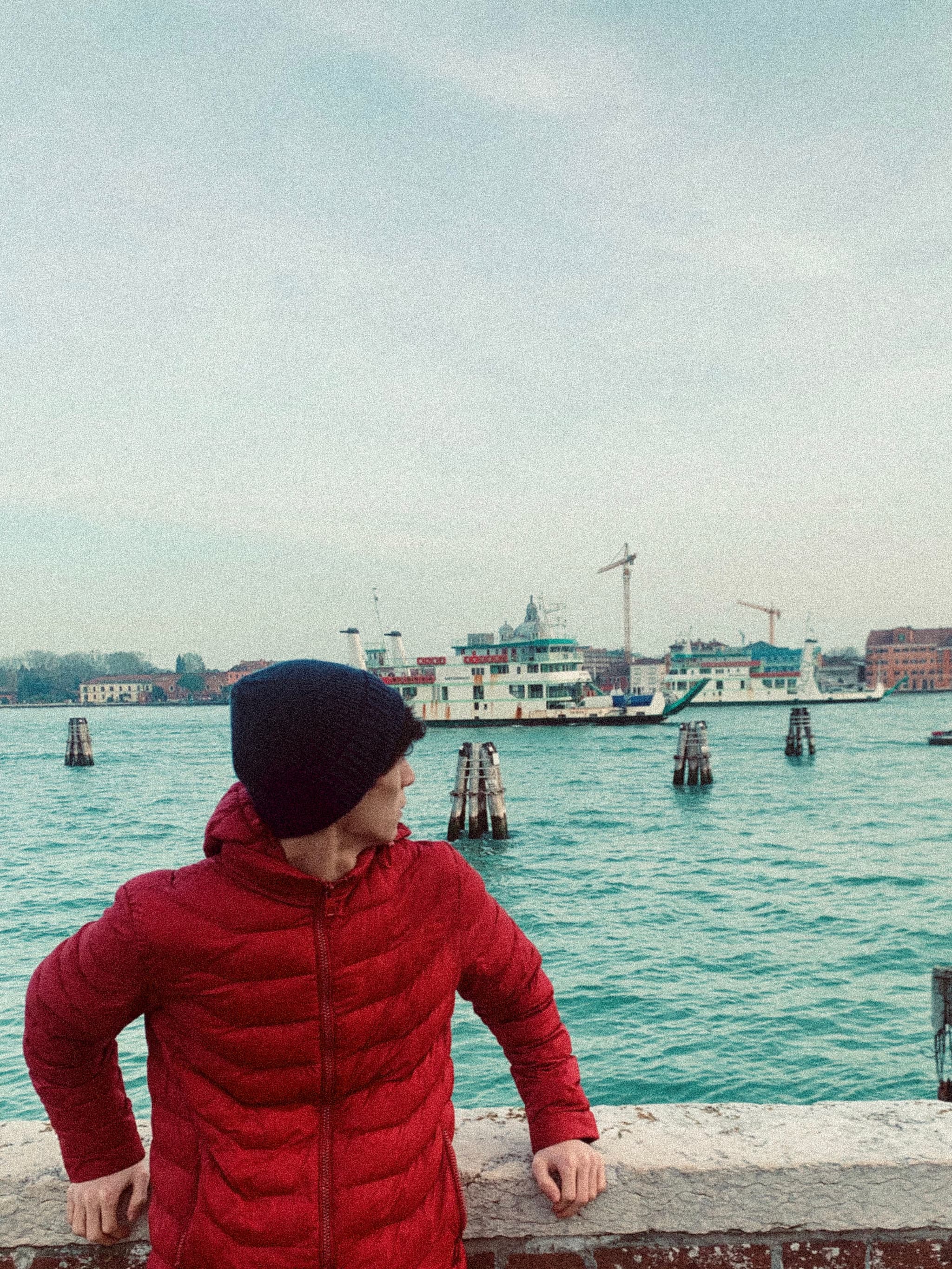 A person in a red jacket stands by the water, gazing at a serene harbor with a distant crane and turquoise sky