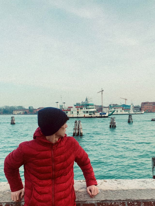 A person in a red jacket stands by the water, gazing at a serene harbor with a distant crane and turquoise sky