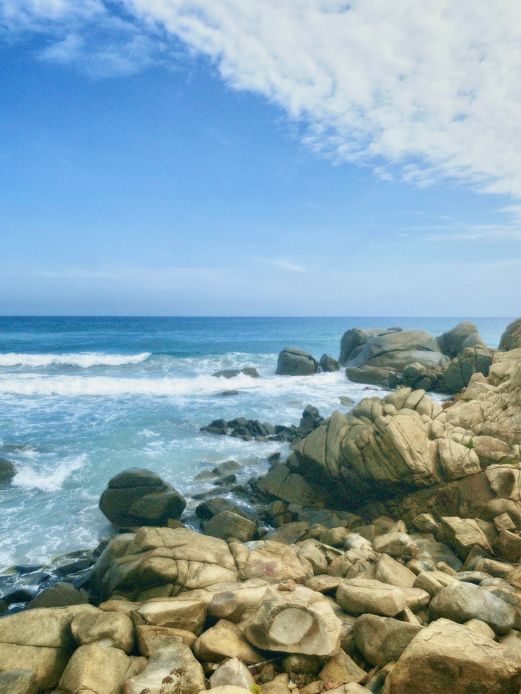 Rocky shoreline under a bright blue sky with white clouds, bathed in natural sunlight, evoking a serene coastal atmosphere
