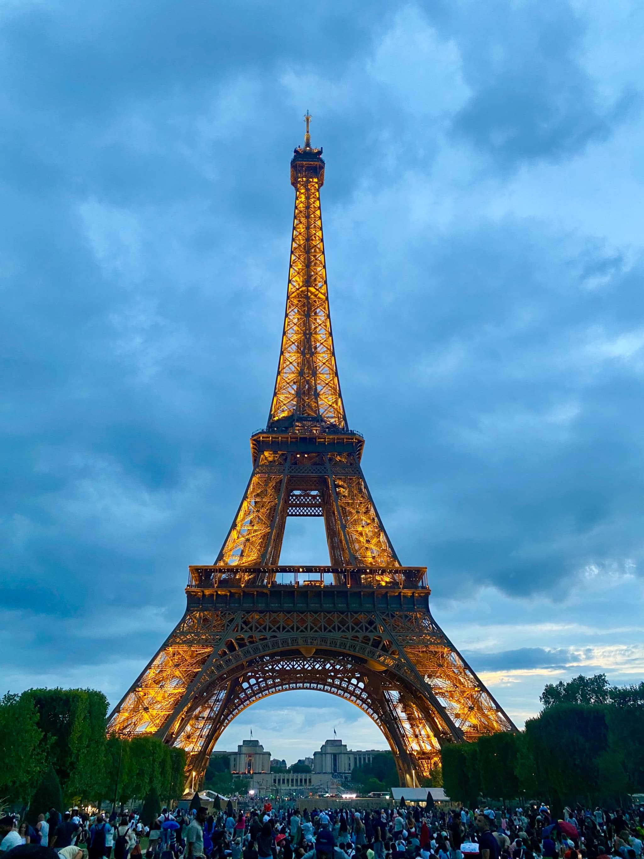 The Eiffel Tower glows warmly under a dramatic, partly cloudy sky, creating a romantic and iconic Parisian scene
