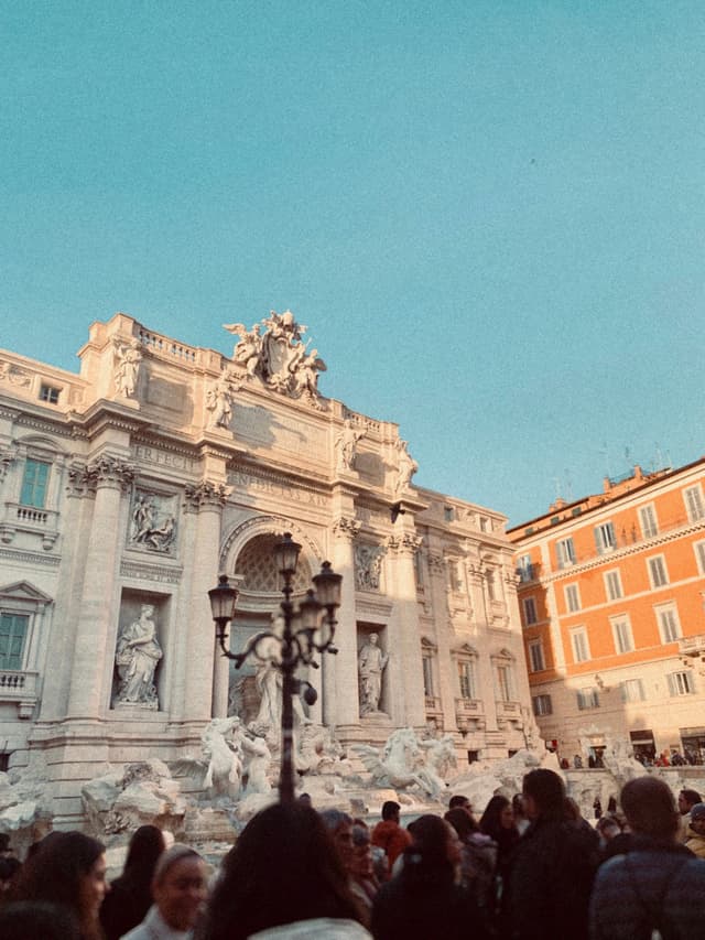 Sunlit Trevi Fountain in Rome, Italy, with crowds in the foreground, clear blue sky, and warm tones