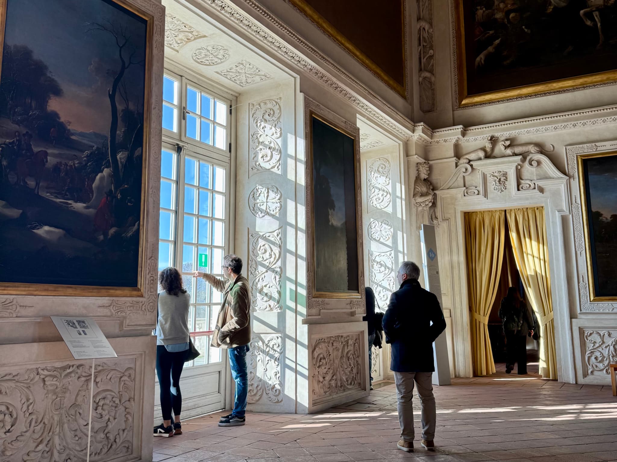 Grand Versailles gallery with people exploring opulent decor, large windows, golden curtains, and rich artwork, bathed in natural sunlight, creating a majestic and elegant atmosphere