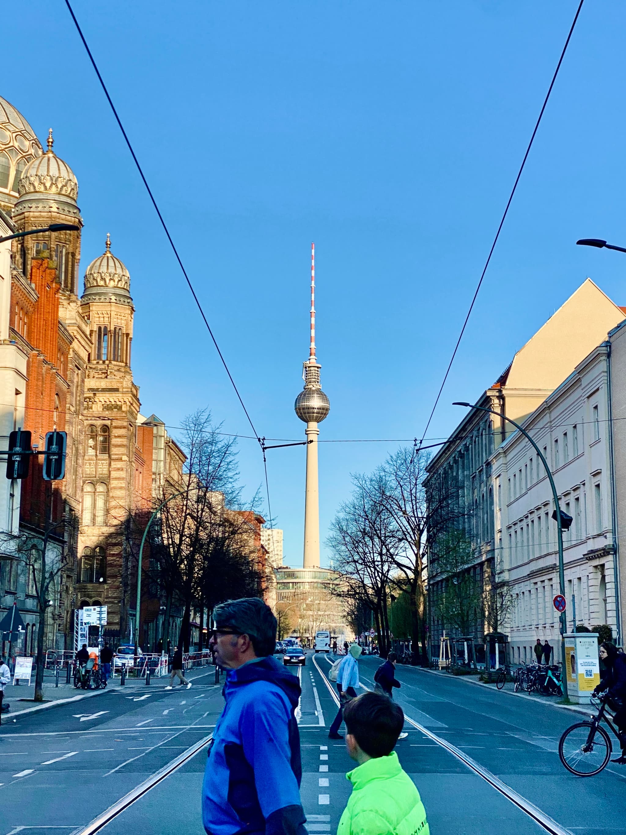 Bright blue sky over Berlin's historic architecture and iconic TV tower, with a cyclist and pedestrian walking under streetlights, capturing urban life in Germany