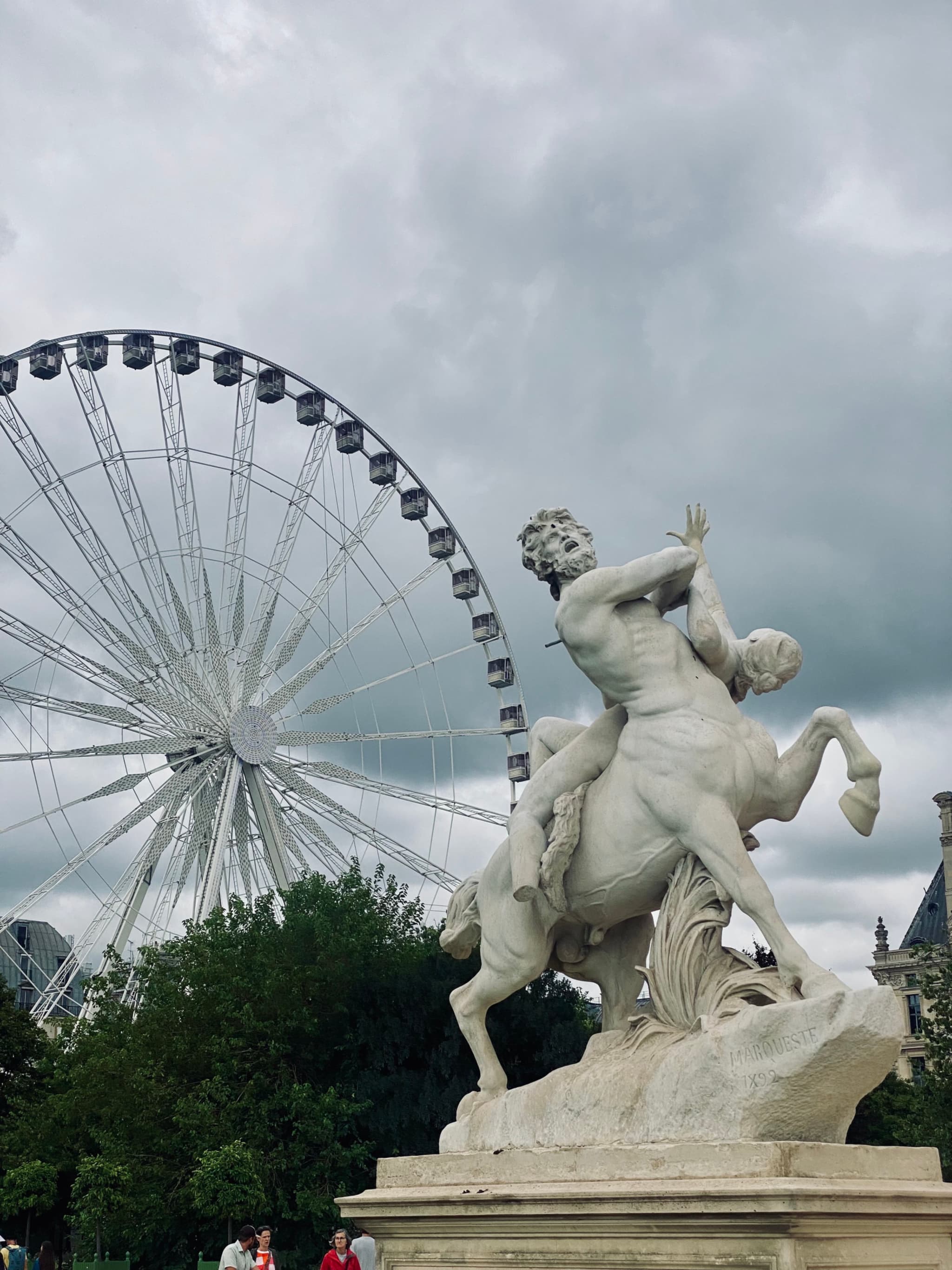 A dramatic sky highlights a statue of a woman on horseback in front of a Ferris wheel, set against the Parisian backdrop of Tuileries Gardens