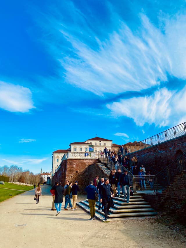 A vibrant daytime scene at a grand, historic castle with people exploring the grand staircase, under a bright blue sky dotted with wispy clouds
